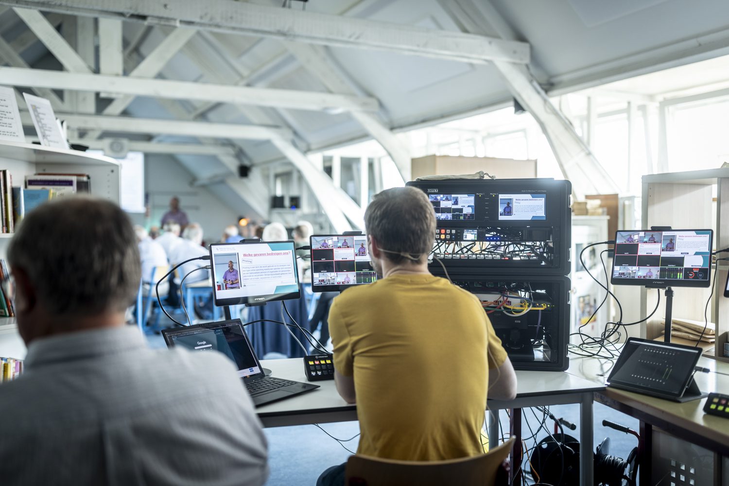 Het clublokaal met de regietafel: schermen met het beeld van de uiteenzetting, en de zaal met leden op de achtergrond.